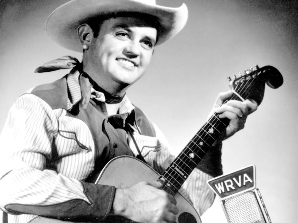 Guitarist Merle Travis, dressed in western clothes, picks an acoustic guitar