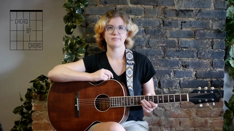 chord by chord instructor kate koenig seated with acoustic guitar in front of a brick background