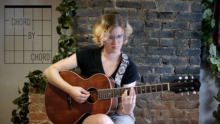 kate koenig, seated playing an acoustic guitar in front of a brick background with ivy