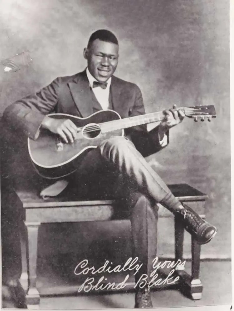 promo photo of Blind Blake seated on a bench playing guitar