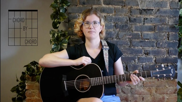 Chord by Chord instructor Kate Koenig, seated with guitar in front of brick backdrop