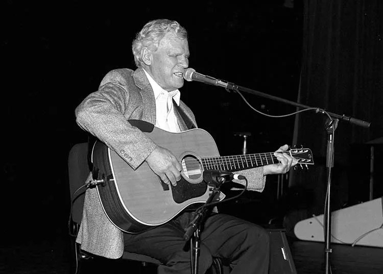 Doc Watson in 1994 playing his guitar and singing into a mic at a concert