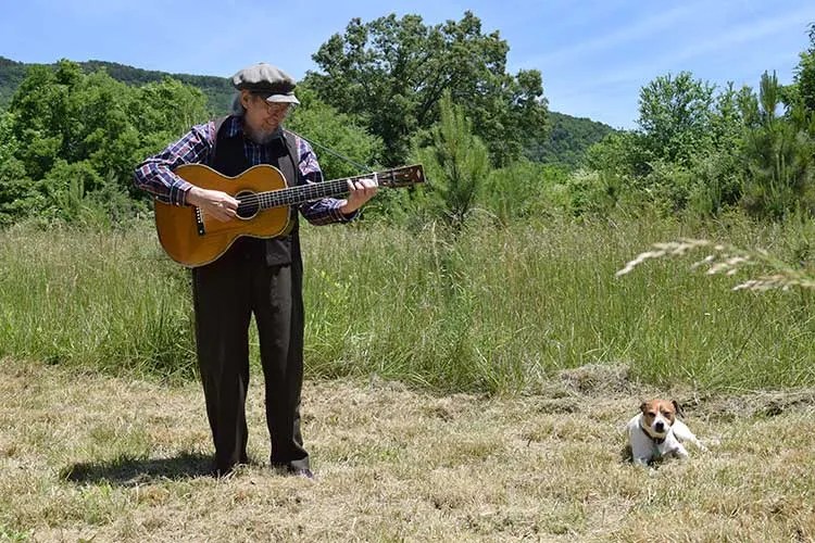 Norman Blake, holding an acoustic guitar, serenades his dog outdoors