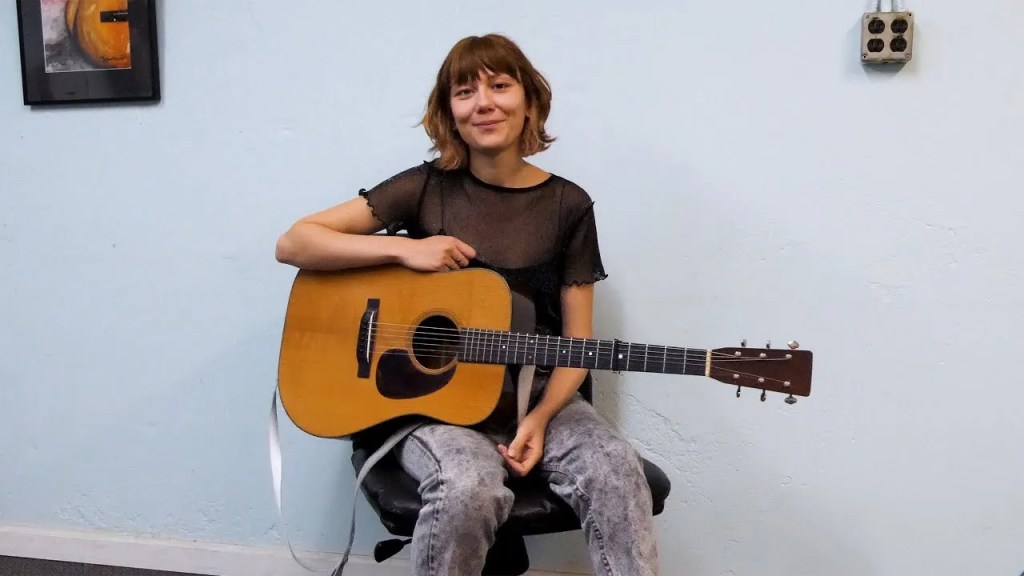 Molly Tuttle smiling with her guitar on a white wall background