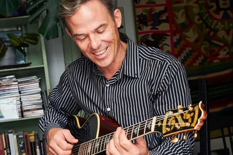 A smiling Greg Ruby seated playing guitar. Photo by Max Branigan