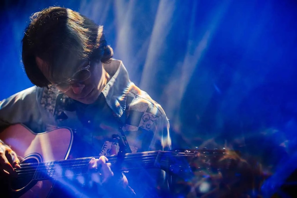 Hayden Pedigo bathed in blue light, playing an acoustic guitar. Photo by Victoria Alexandrova