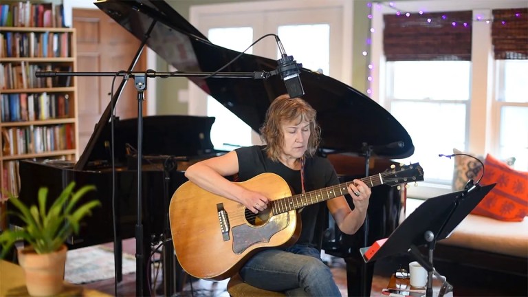 Jane Miller seated teaching a weekly workout lesson on her acoustic guitar, photographed in a studio with a microphone in front and a grand piano in the background.