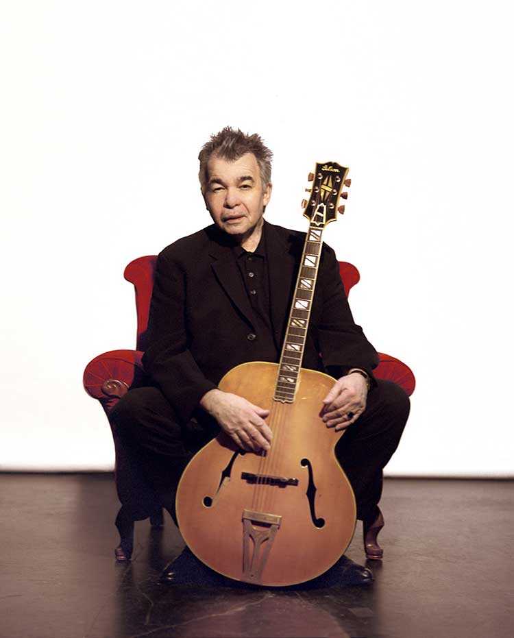 guitarist john prine sitting in a chair with guitar