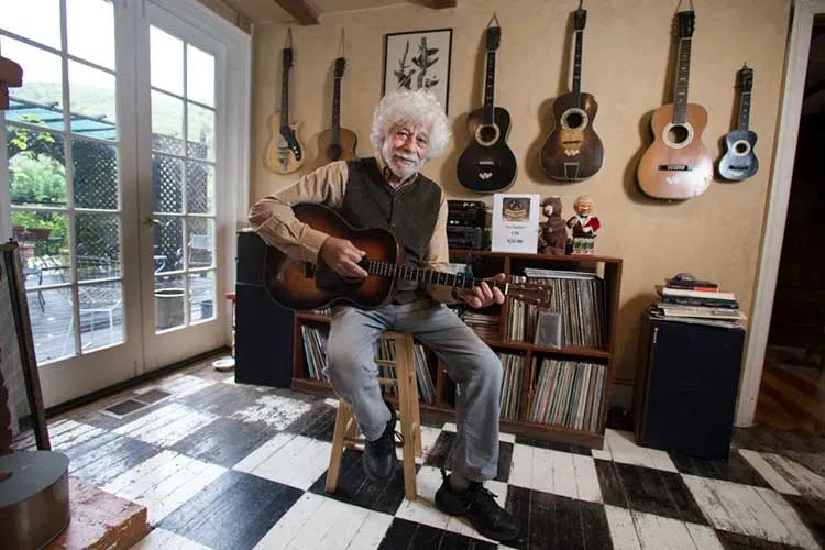 Lowell Levinger seated playing a tenor guitar in front of a wall of other guitars.