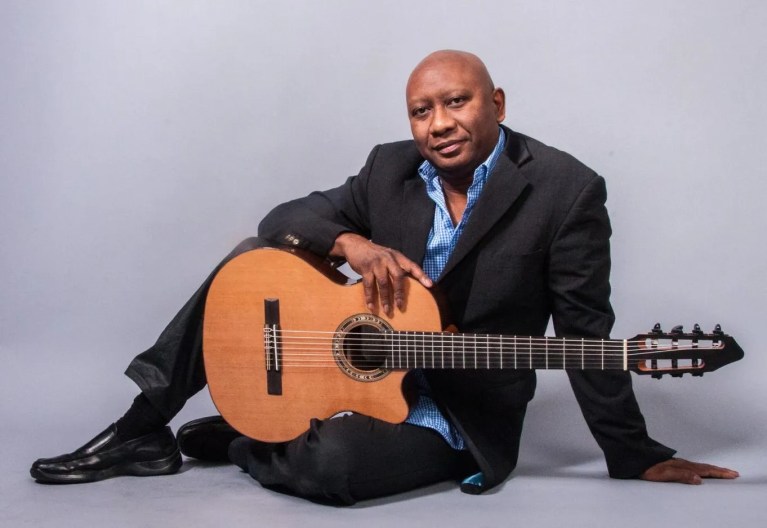 Photograph of Ron Jackson wearing a suit, seated on the floor with his acoustic guitar balanced across one knee.