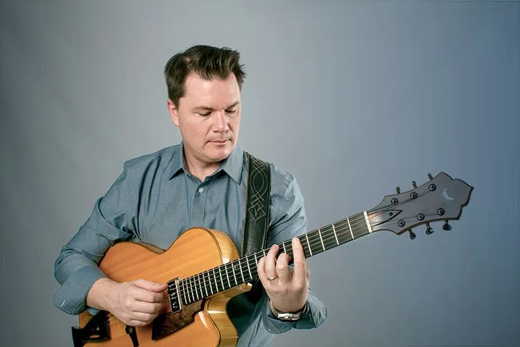 sean mcgowan wearing chambray shirt with gray studio backdrop, demonstrating lesson material on his acoustic guitar