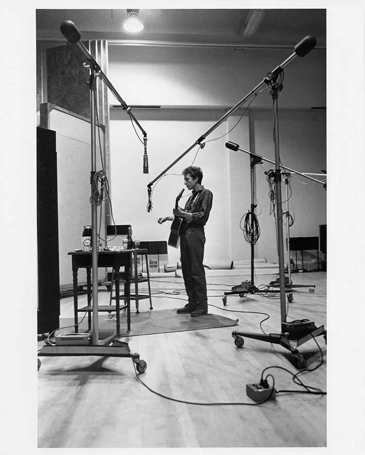 Bob Dylan stands alone in a recording studio with an acoustic guitar