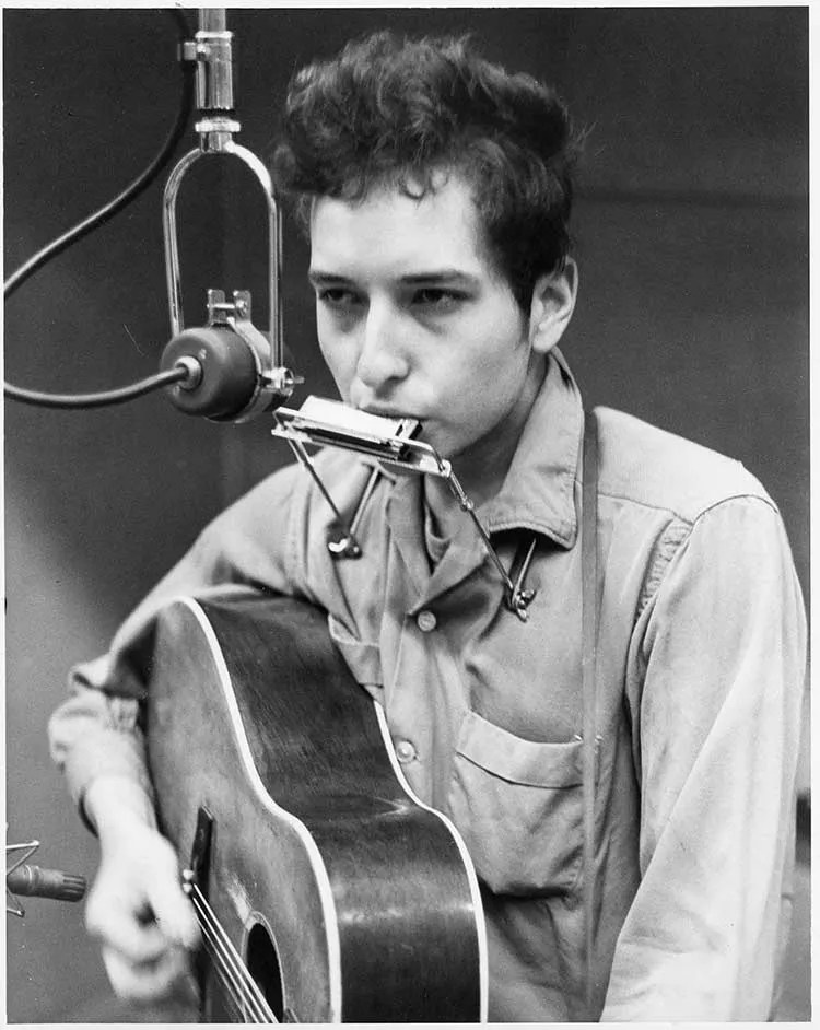 bob dylan seated in front of a microphone playing harmonica and acoustic guitar