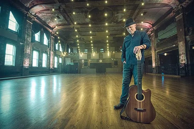 Willy Porter standing with guitar in an empty concert hall. Photo by Cory Zimmerman/Z2