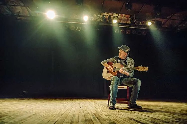 Willy Porter seated with his nine-string guitar on an empty stage. Photo by Cory Zimmerman/Z2