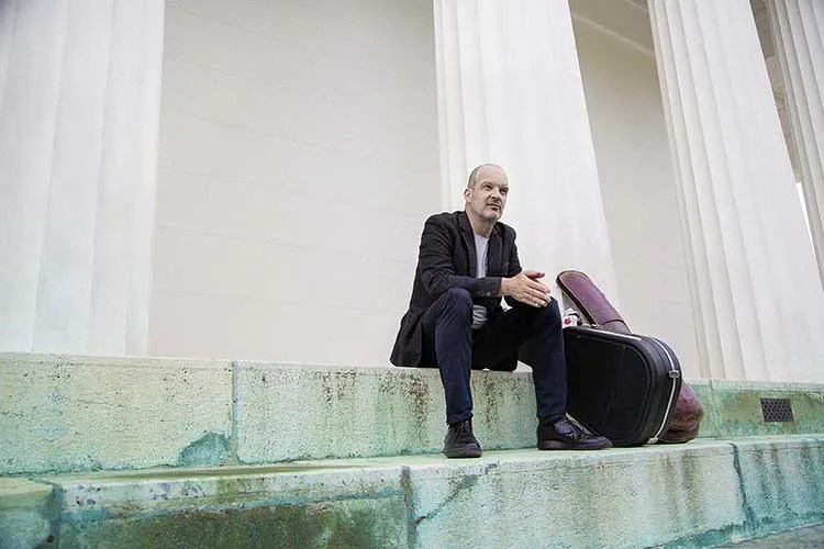 Wolfgang Muthspiel seated on steps beneath columns net to guitar cases. ©-Laura-Pleifer